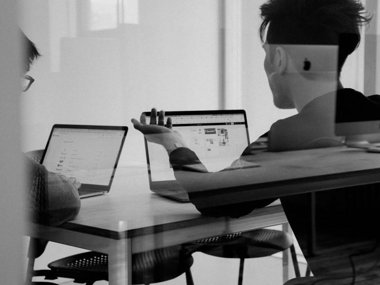 Two people sitting at a table working on laptops, discussing ideas in a modern workspace.