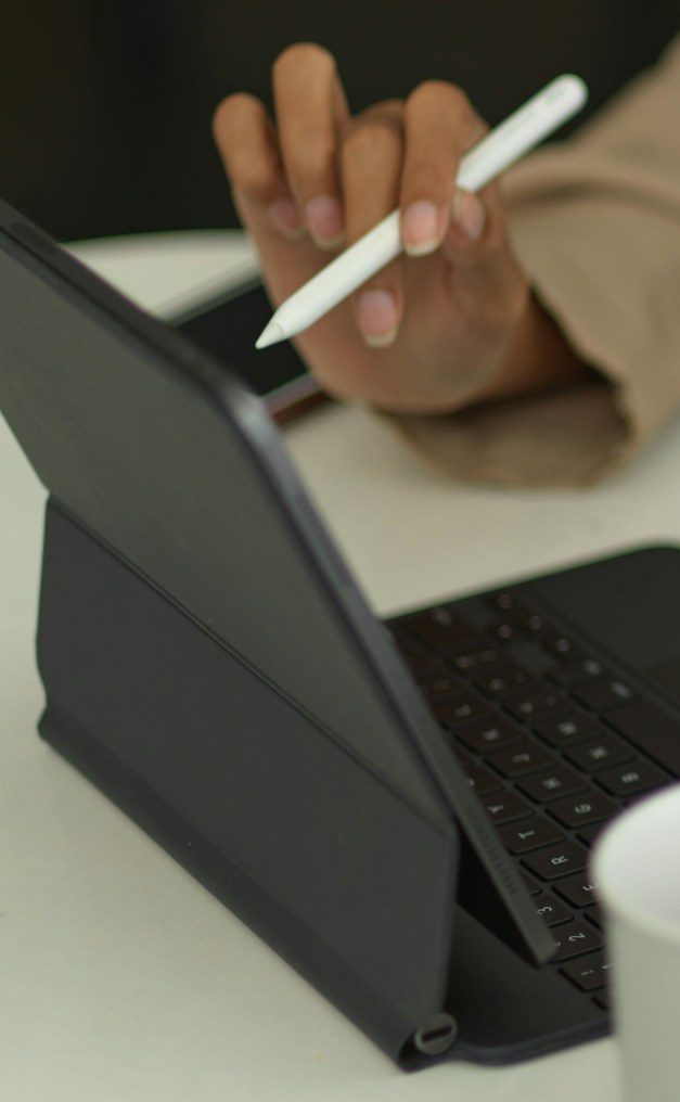 A hand using a stylus on a tablet beside a cup and keyboard.