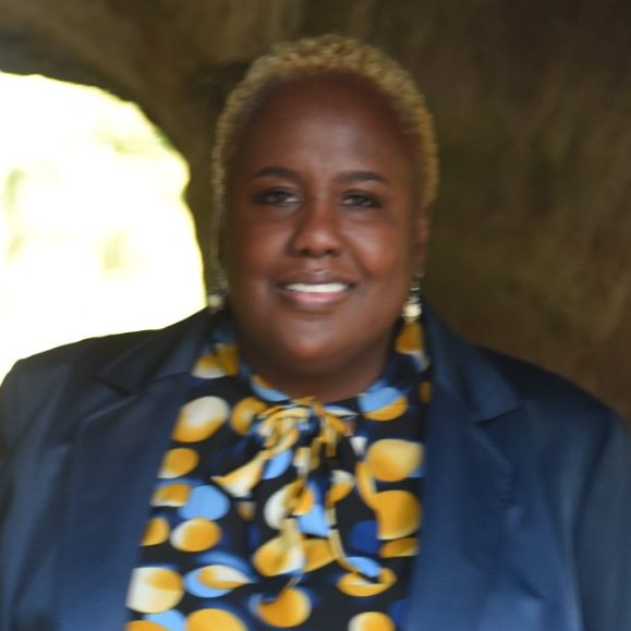 Smiling woman with short, curly blonde hair wearing a dark blazer and patterned scarf.