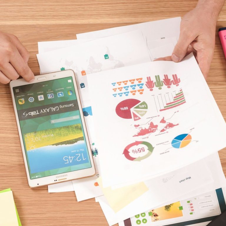 Hands holding a tablet and a printed report with colorful charts and graphs on a wooden table.